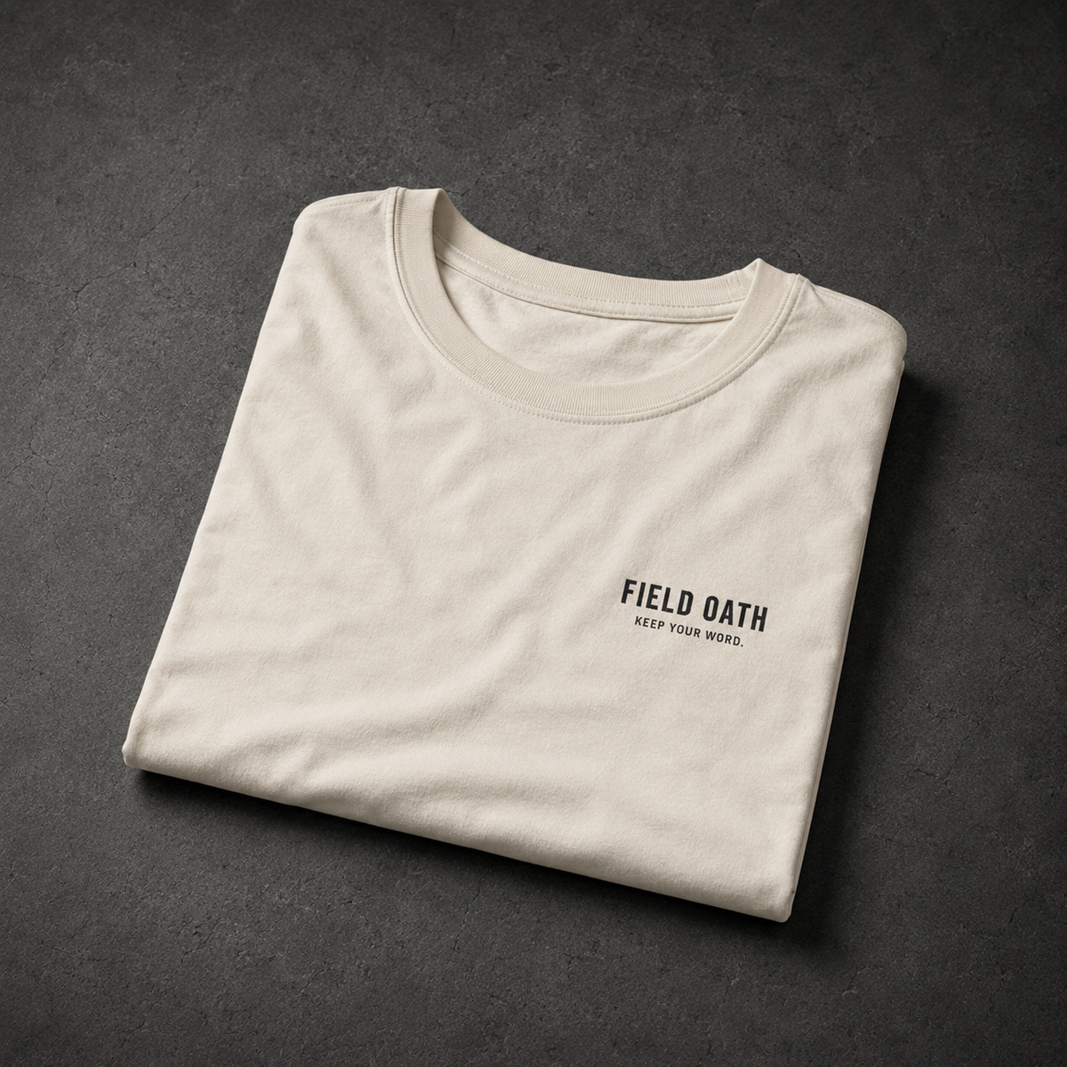 Bone FIELD OATH Standard Mark T-Shirt folded on a dark concrete studio surface